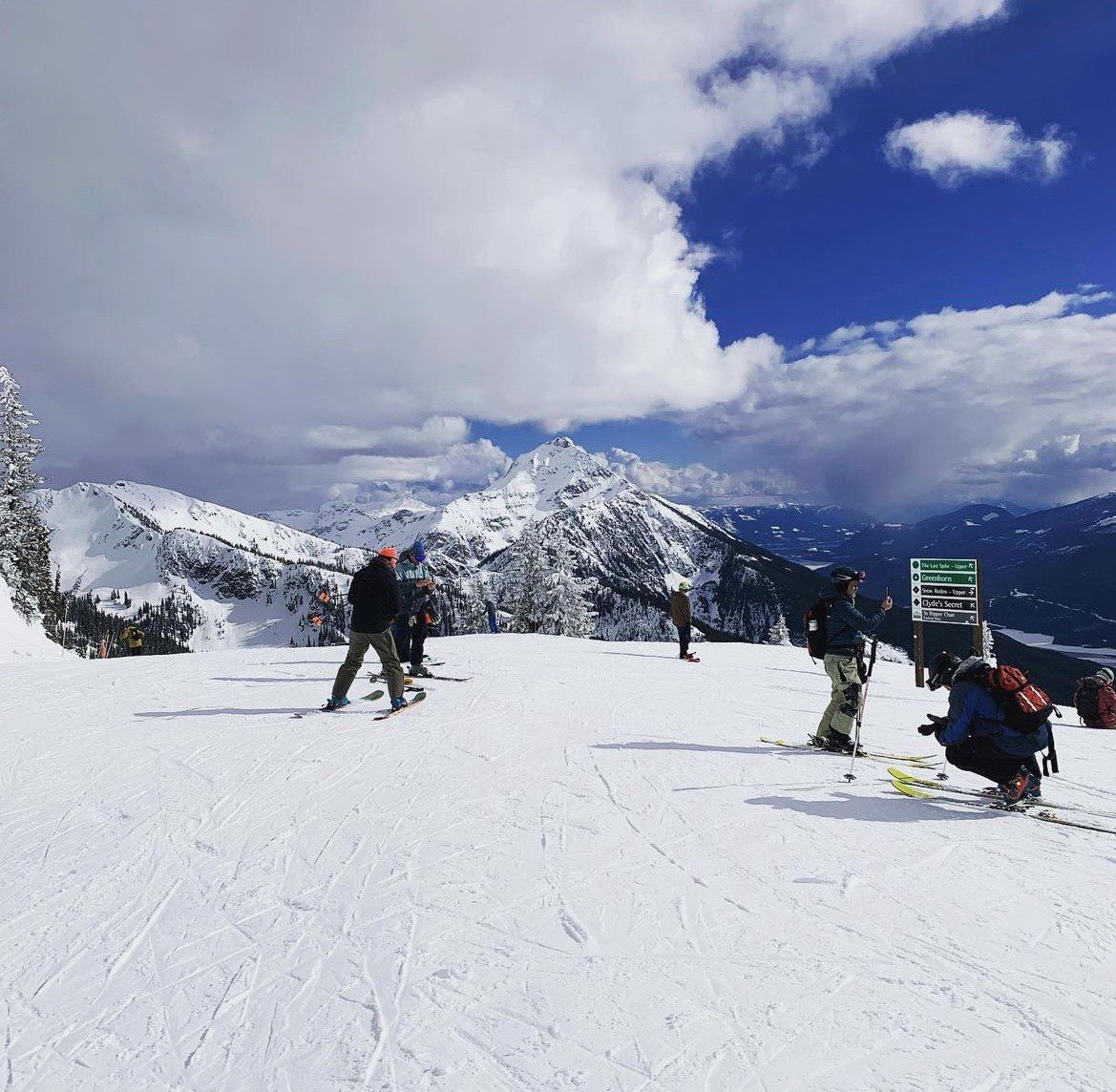 Several skiers and snowboarders on a snow-covered mountain with cloudy sky and distant peaks in the background.