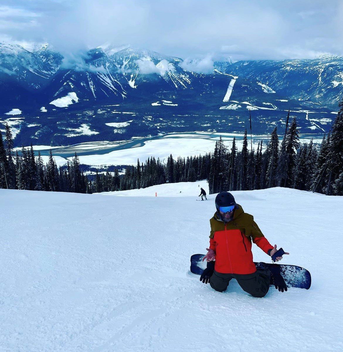 Snowboarder kneeling on a snowy slope with a scenic mountain and forest backdrop, wearing a red and green jacket and helmet. Another person is snowboarding downhill in the distance.