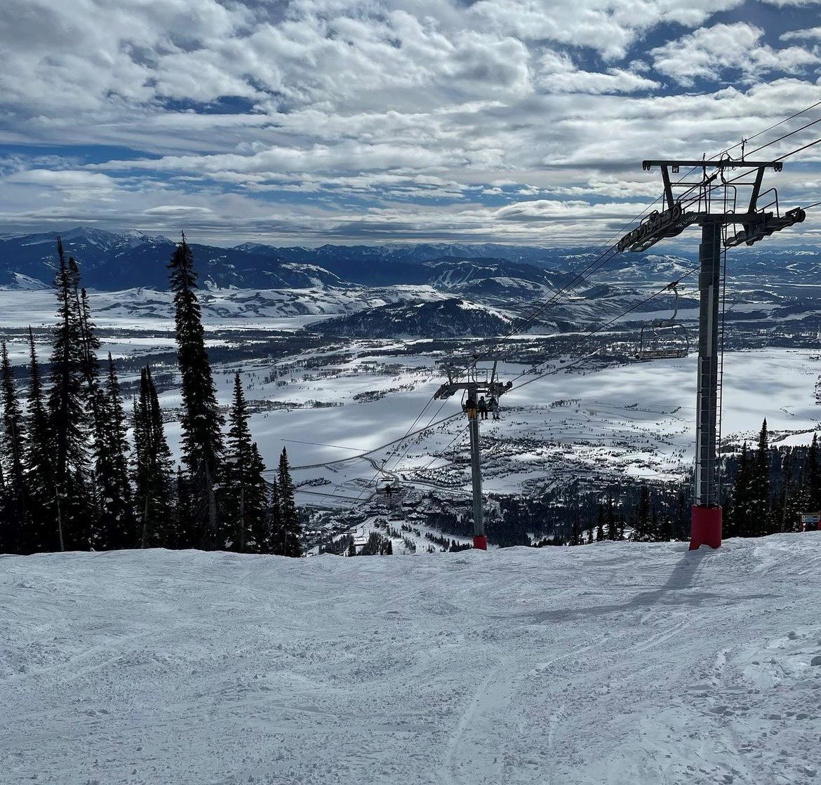 Snow-covered landscape with a ski lift and distant town in the valley. Tall trees on the slope, mountains visible on the horizon under a partly cloudy sky.